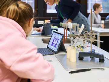 Schülerinnen sitzen in einem Physiklabor und schauen in Zweiergruppen auf Tablet-Bildschirme.