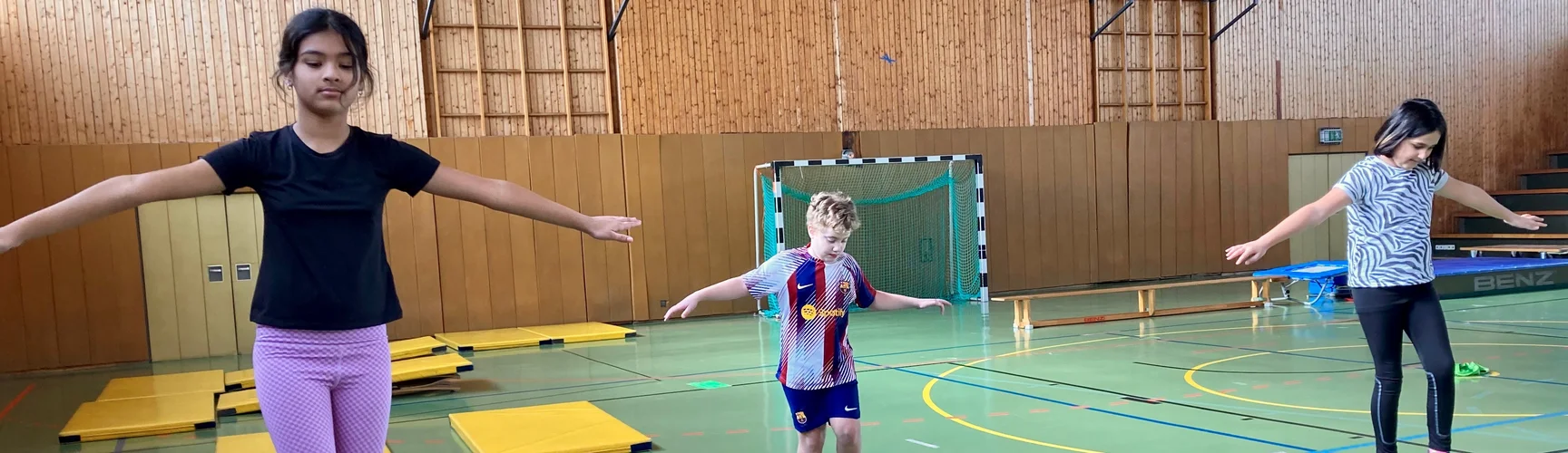 In einer Sporthalle balancieren drei Kinder auf verschiedenen Wegen: Auf einem Balken, der auf Kästen liegt, auf einem bunten Steg mit vielen Kurven und auf einer umgedrehten Bank.