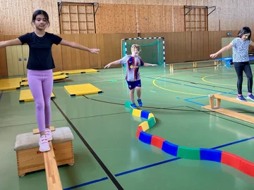In einer Sporthalle balancieren drei Kinder auf verschiedenen Wegen: Auf einem Balken, der auf Kästen liegt, auf einem bunten Steg mit vielen Kurven und auf einer umgedrehten Bank.