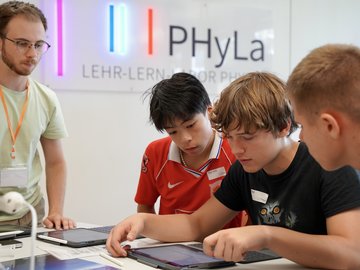 Eine Lehrkraft betreuut drei Schüler, die gemeinsam auf einen Tablet-Bildschirm schauen.