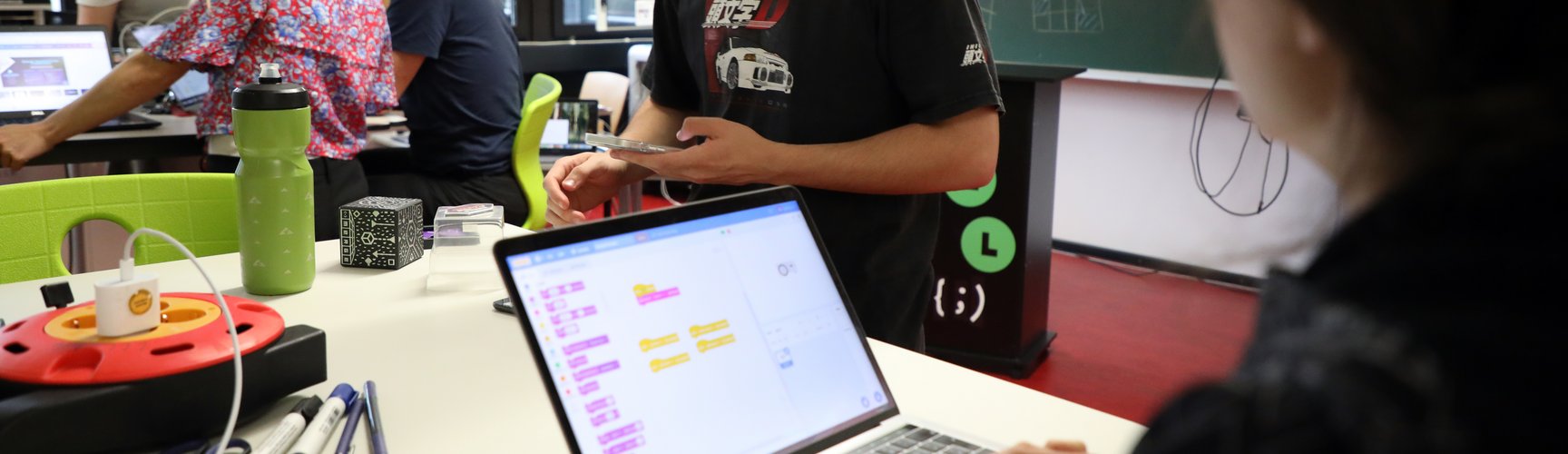 Im Lehr-Lern-Labor Informatik mit Stehtischen und grünen Stühlen sind Menschen an den Tischen mit ihren Smartphones und Laptokps beschäftigt. Ein junger Mensch hält einen Würfel in der Hand.