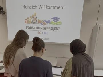 Drei Schülerinnen sind von hinten zu sehen, wie sie gemeinsam eine Forshcungsfrage entwicklen. Eine trägt ein Kopftuch.