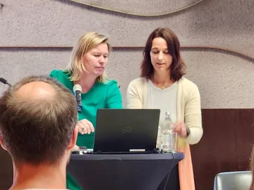 Zwei Frauen stehen an einem Rednerpult mit Laptop und Mikrofon, im Vordergrund ist der Hinterkopf eines Zuhörers zu sehen.