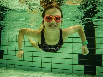 Ein Mädchen mit Schwimmbrille schwimmt in einem Schwimmbecken unter Wasser, es lächelt.