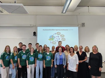 Beim Austauschtreffen Sportschuhbande posiert eine Gruppe in einem Seminarraum für das Foto, einige tragen ein grünes T-Shirt mit dem Logo der Sportschuhbande.
