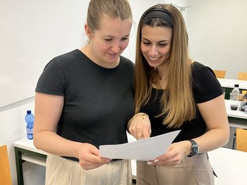 Zwei Studentinnen stehen in einem Seminarraum und betrachten gemeinsam ein Arbeitsblatt.
