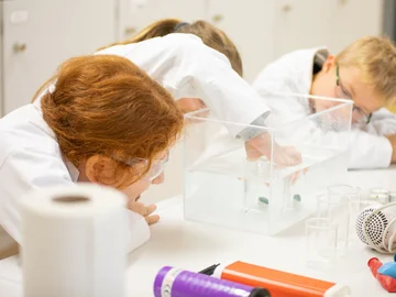 Drei Kinder experimentieren mit einem Gegenstand in einem wassergefüllten Behälter, sie tragen Laborkittel und Schutzbrillen.