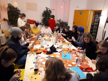 Eine Gruppe Studierender sitzt um einen Tisch und bastelt, auf dem Tisch liegen viele Materialien wie Papiere und Nüsse.