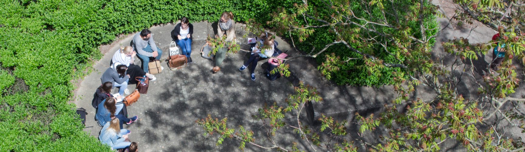 Eine Gruppe Personen sitzt auf dem Campus in einem durch eine Hecke geschützten Bereich