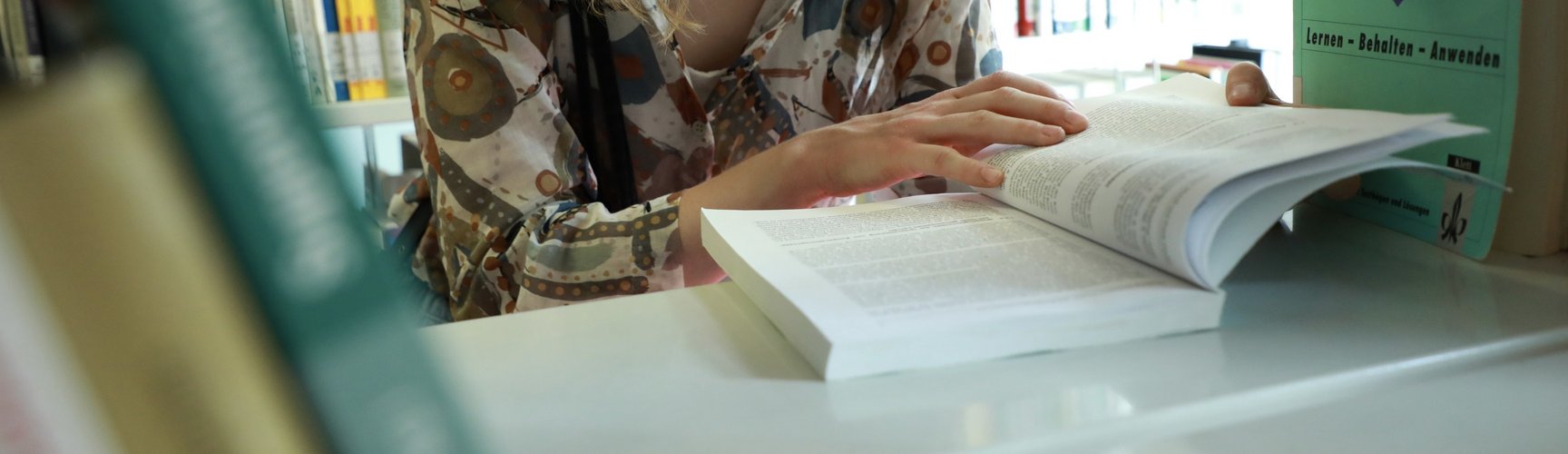 Eine junge Frau mit Brille blickt in der Bibliothek in ein Buch an einem Bücherregal.