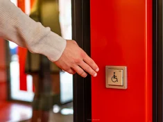 eine Hand ist dabei, auf einen automatischen Türöffner eines barrierefreien Eingangs zu drücken
