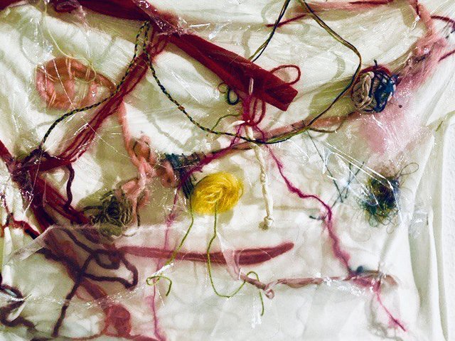 Ein abstraktes Bild zeigt ein Geflecht aus bunten Fäden und Garnen, das unter und auf einer zerknitterten, transparenten Kunststofffolie liegt. Besonders auffällig ist ein kleines, leuchtend gelbes Garnknäuel in der Mitte, umgeben von roten, rosa und dunkleren Fäden.