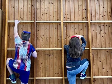 Zwei Kinder klettern an einem Klettergerüst aus Holz nach oben, sie balancieren jeweils ein kleines Säckchen auf dem Kopf.