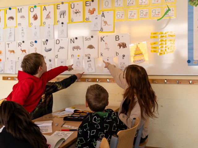 Vie Kinder im Klassenzimmer zeigen mit ihren Fingern auf eine Pinnwand mit Bildern von Tieren und deren Anfangsbuchstaben