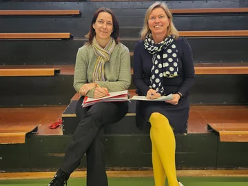 Zwei Frauen sitzen in einer Sporthalle auf einer kleinen Tribüne.