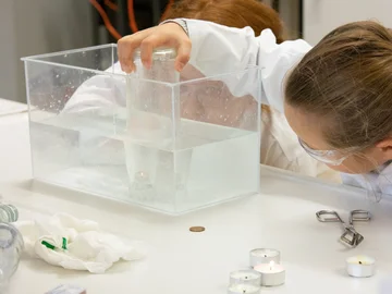Zwei Kinder in Laborkitteln beobachten und experimentieren mit Teelichtern, die in einem Wassertank unter Wasser stehen, geschützt durch eine Flasche.