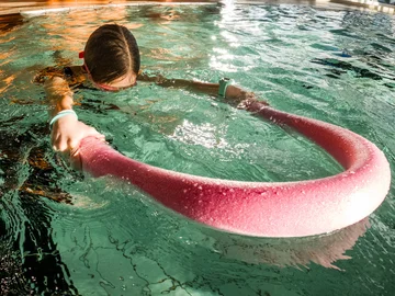 Ein Kind schwimmt in einem Schwimmbecken, es hat das Gesicht unter Wasser und hält eine Schwimmnudel vor sich.
