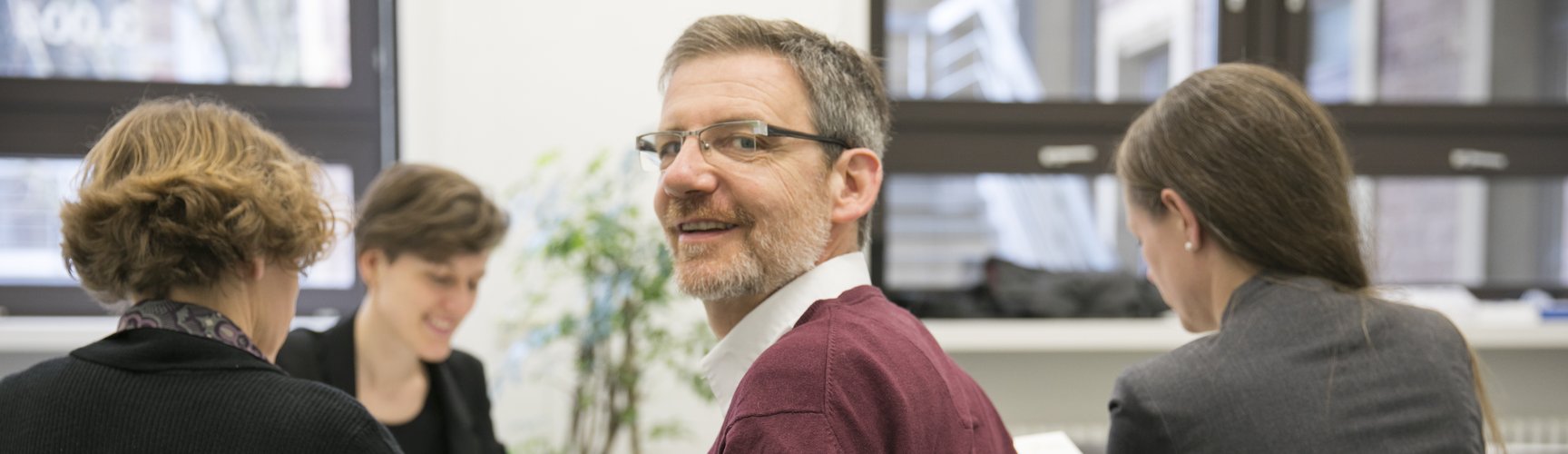 Drei Frauen und ein Mann sitzen um einen Besprechungstisch mit Unterlagen, der Mann dreht sich zur Kamera um und lächelt.