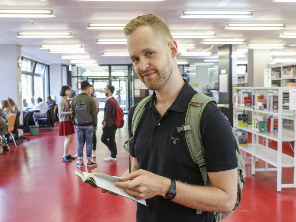 Ein Student, von vorne fotografiert, liest ein Buch in der Bibliothek im Gebäude 2 der PH Karlsruhe. Hinter ihm sind mehrere Studierende zu sehen. ©PHKA