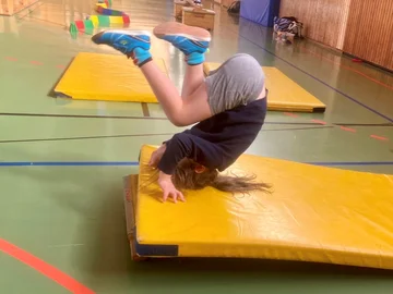 In einer Sporthalle führt ein Kind eine Rolle vorwärts auf einer leicht erhöhten Bodenmatte aus.