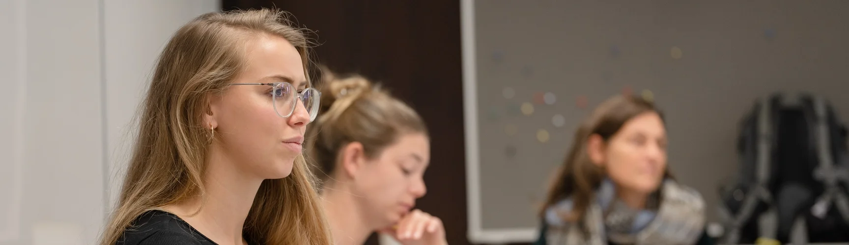 Drei Studentinnen sitzen in einem Seminarraum und hören zu, eine macht sich Notizen.