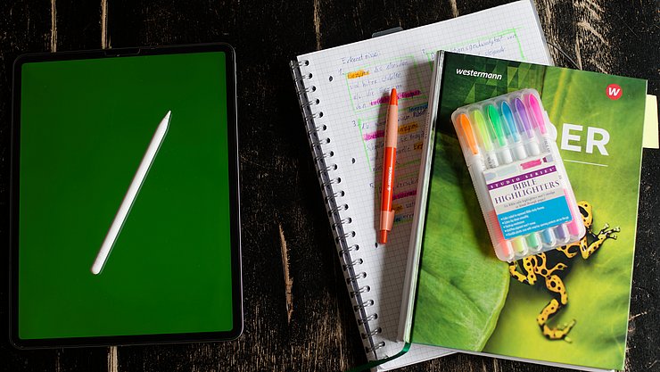 Symbolbild KI in der Schule Ein Tablet und ein Schulbuch liegen symbolisch nebeneinander. Das Schubuch liegt zudem auf einem Block und es gibt einige bunte Stiftte.