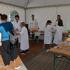 PHKA Chemie bei EFFEKTE 2023 Blick in das Pagodenzelt des Lehr-Lern-Labors "Kinder experimentieren" bei EFFEKTE 2023 - auf Biertischen in einem U sind Experimente aufgebaut, dazwischen Kinder und Studierende in weißen Laborkitteln und mit Schutzbrillen.