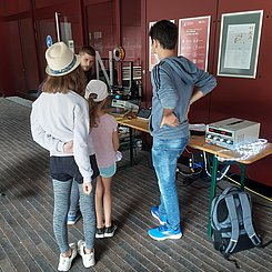 Es sind drei Kinder und Jugendliche von hinten im Freien an einem Tisch an einer roten Wand zu sehen, worauf naturwissenschaftliche Experimente aufgebaut sind, darüber sind Plakate. Ein Student erklärt das Experiment.