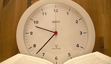 2022-07-15_Öffnungszeiten-Bibliothek Wanduhr mit einem aufgeschlagenen Buch davor