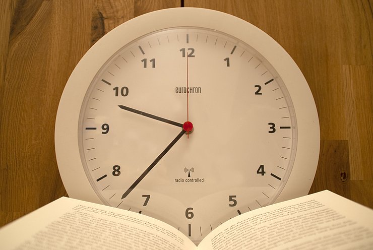 2022-07-15_Öffnungszeiten-Bibliothek Wanduhr mit einem aufgeschlagenen Buch davor