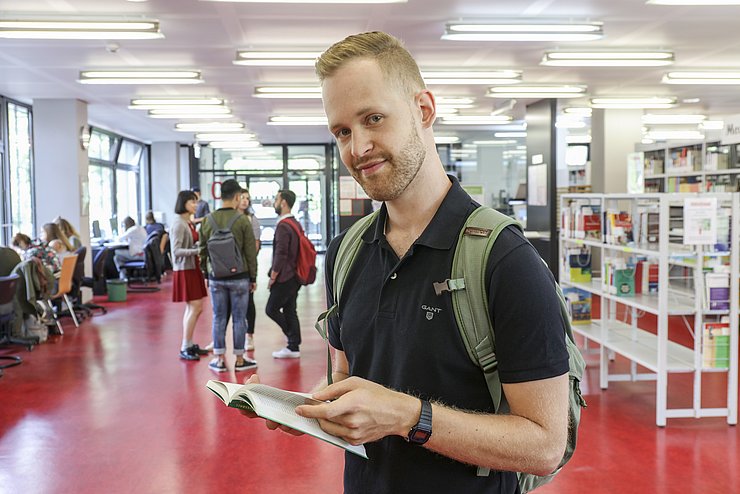 Ein Student, von vorne fotografiert, liest ein Buch in der Bibliothek im Gebäude 2 der PH Karlsruhe. Hinter ihm sind mehrere Studierende zu sehen. ©PHKA
