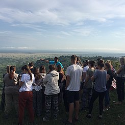 Das Bild zeigt die praktische Arbeit im Feld der Geographie. Zusehen ist eine Gruppe von Studierenden, die die umliegende Landschaft betrachten und einzelne Besonderheiten vom Dozenten im Feld durch Zeigen und Erklärungen erläutert bekommen.
