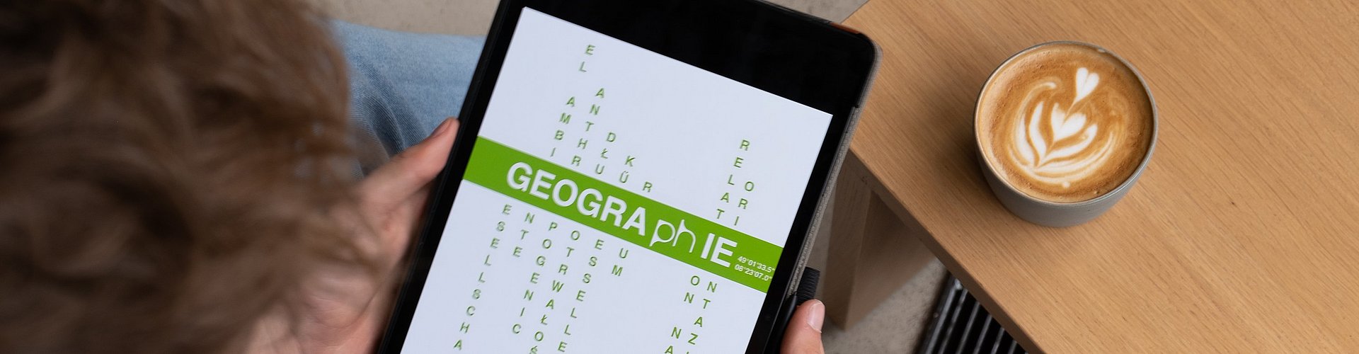 Es wird ein Tablet mit dem Logo des Faches gezeigt. Das Logo zeigt die Bedeutung von Geographie an der Pädagogischen Hochschule Karlsruhe, wie Raum mehrsprachig zwischen Gesellschaft und Umwelt verhandeln.