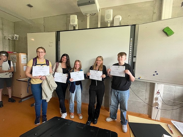 Zertifikate EntreCo Think Hackathons Fünf jugendliche Schüler:innen zeigen stolz ihre durch einen Hackathon des Projekts EntreCo Think erworbenen Zertifikate vor einem Whiteboard.