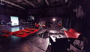 Das Bild zeigt einen großen, dunklen Studio- oder Theaterraum mit hoher Decke. Auf dem Boden stehen verteilt Kamera- und Lichtstative sowie viele Kabel. In der Mitte liegt ein roter Teppich mit runden Sitzkissen und kleinen Hockern. Links hinten ist ein grüner Hintergrund (Greenscreen) mit Stühlen und Technik davor. Rechts im Vordergrund stehen ein rotes Sofa, ein schwarzer Sessel und ein kleiner Tisch. Der Raum wirkt wie eine provisorische Bühne oder ein kreatives Setup ohne Menschen.
