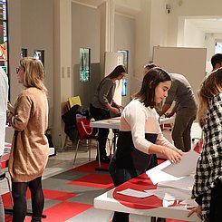 Studierende und Schülerinnne und Schüler stehen an verschiedenen Tischen in einem hellen Raum am Museum am Markt und entwerfen gemeinsam Ökonomische Lernspiele. REchts ist Prof. Dr. Claudia wiepcke im Profil zu sehen.nsam 