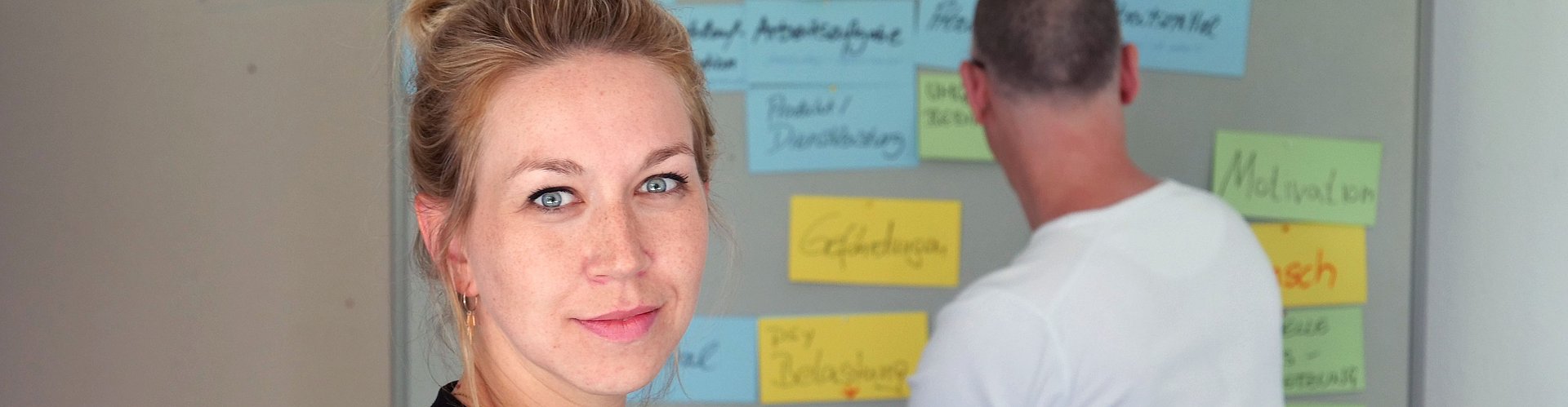 eine Person steht im Vordergrund und blickt in die kamera. Eine Person im Hintergrund arbeitet an einer Mindmap.