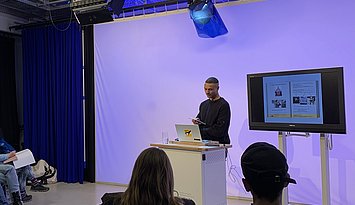 Eine Person hält einen Vortrag an einem Rednerpult in einem Studio, während mehrere Zuhörende sitzen und auf einen Bildschirm mit Präsentationsfolien schauen.